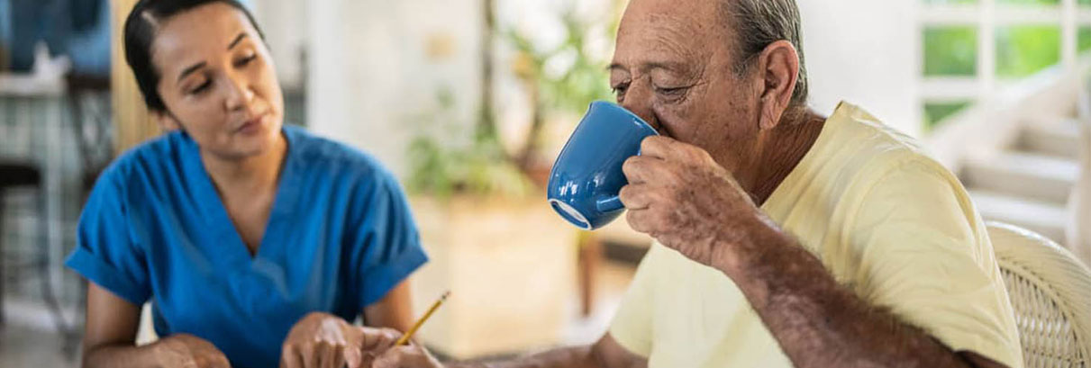 Caregiver and client sitting at a table in a sunny kitchen while client drinks a cup of coffee 
